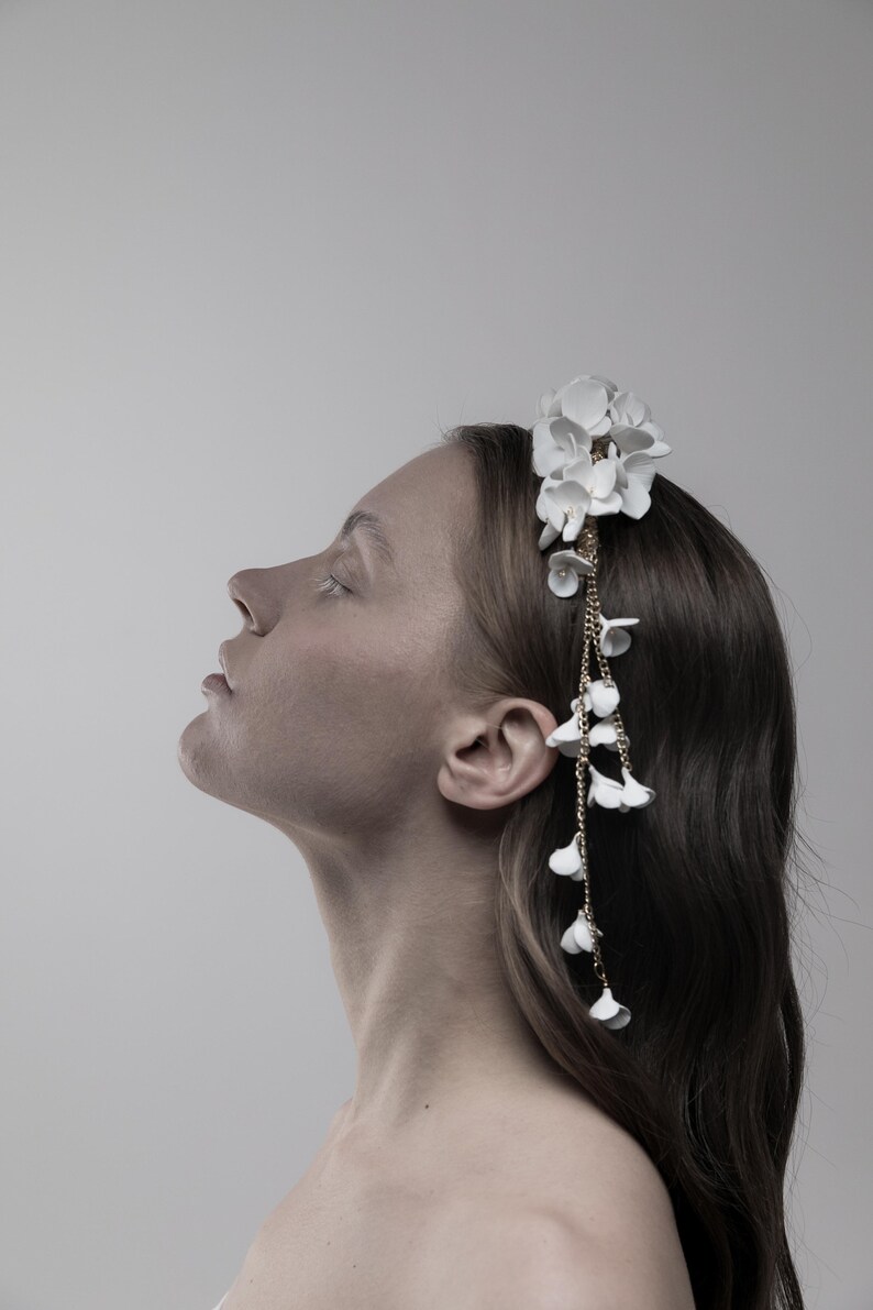 May include: A white floral headband with a gold chain and white flower accents. The headband is worn by a person with long brown hair.
