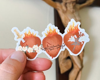 May include: Three red, flaming hearts with thorns and white flowers. The hearts are arranged horizontally and are surrounded by a white background.