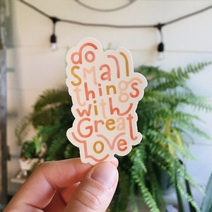 Puede incluir: Una pegatina con el texto "Do small things with great love" en una fuente estilizada y colorida. La pegatina es de color melocotón, rosa y dorado.