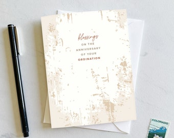 May include: A white card with a brown textured background. The text "blessings on the anniversary of your ordination" is printed in red on the card.