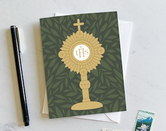 May include: A green card with a gold monogram of the letters "IHS" surrounded by a sunburst design. The monogram is on a pedestal with a cross at the top. The background is a green leafy pattern.