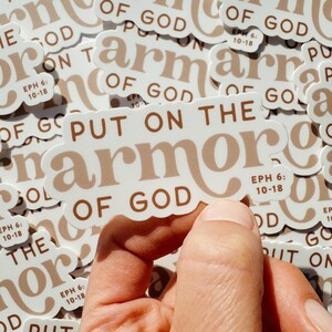 May include: A white sticker with brown text that reads "Put on the armour of God" and "Eph 6:10-18". The sticker is being held by a hand.