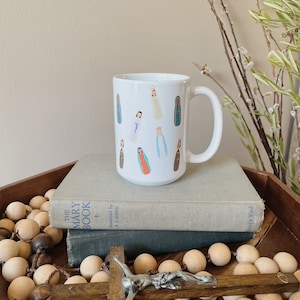 May include: White ceramic mug with a colorful illustration of various depictions of Mary, the mother of Jesus. The mug is sitting on top of two books.