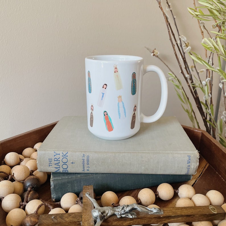 May include: White ceramic mug with a colorful illustration of various depictions of Mary, the mother of Jesus. The mug is sitting on top of two books.