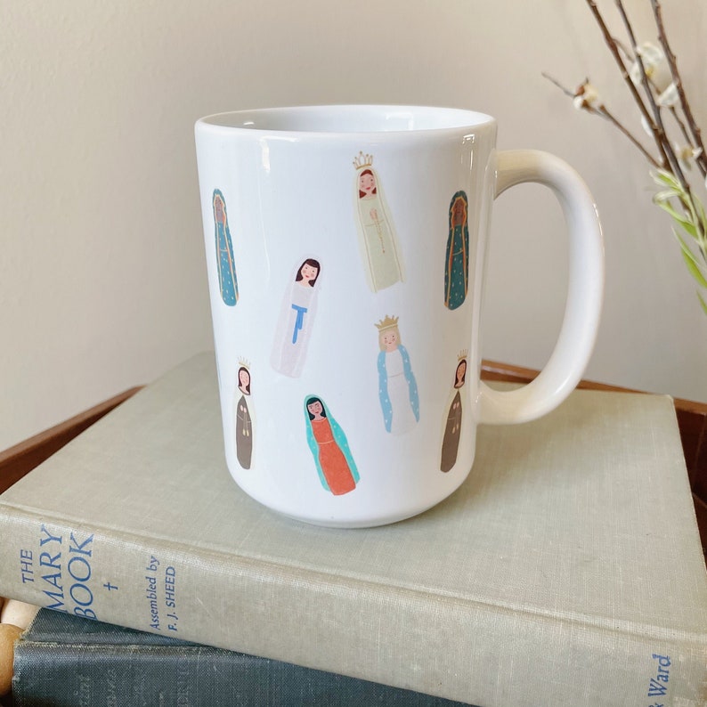 May include: White ceramic mug with a colorful pattern of various depictions of the Virgin Mary. The mug has a handle on the right side.