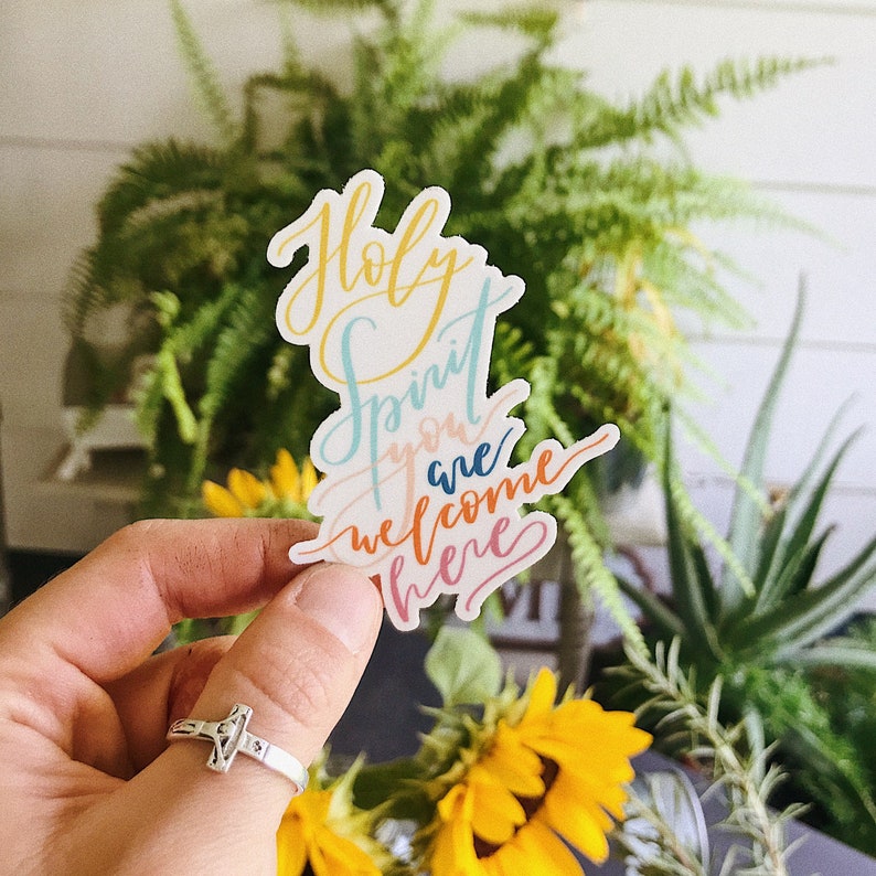 K&ouml;nnte beinhalten: Ein Aufkleber mit den Worten "Holy Spirit you are welcome here", geschrieben in bunten Kursivbuchstaben. Der Aufkleber wird von einer Hand mit einem silbernen Ring gehalten.