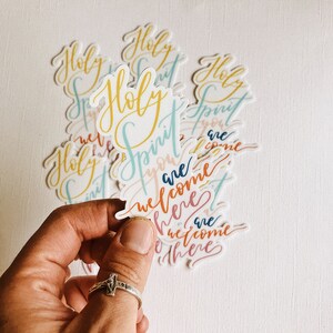 K&ouml;nnte beinhalten: Ein Satz bunter Aufkleber mit dem Schriftzug "Holy Spirit you are welcome here" in einer kursivschriftlichen Schrift.