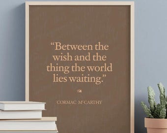 May include: A framed print featuring the quote "Between the wish and the thing the world lies waiting." by Cormac McCarthy. The text is light brown on a dark brown background. A stack of books and a potted plant are in the foreground.