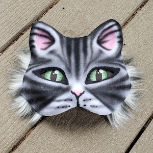 May include: A gray and white cat mask with green eyes and a pink nose. The mask has a furry trim around the edges.