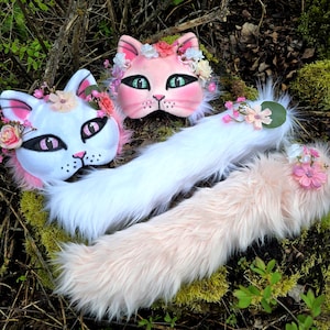 May include: Two cat masks and two fluffy tails. One mask is white with pink accents, the other is pink. Both have floral headbands and cat-eye makeup. One tail is white, the other is light pink, both are fluffy.