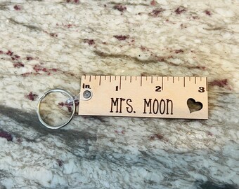 May include: Wooden ruler keychain with a heart cutout and the text "MRS. MOON" engraved on it. The keychain is attached to a silver ring.