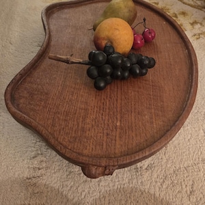 May include: A wooden serving tray with a unique, curved shape. The tray is filled with a pear, an orange, a bunch of grapes, and a few red cherries. The wood has a warm, brown tone, and the tray has a decorative edge.