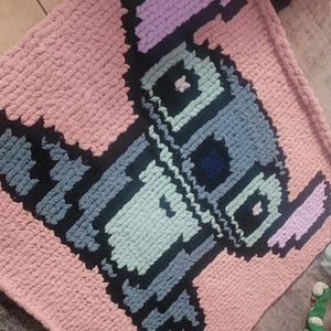 May include: A handmade, pink, pixelated blanket featuring a cartoon character design in shades of blue, black, and purple. The blanket is made of a soft, chunky yarn, perfect for a baby or child's room.