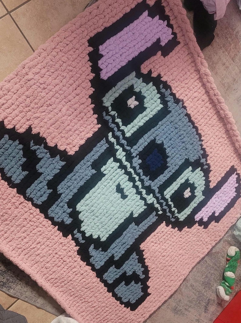 May include: A handmade, pink, pixelated blanket featuring a cartoon character design in shades of blue, black, and purple. The blanket is made of a soft, chunky yarn, perfect for a baby or child's room.