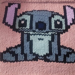 May include: A pink blanket with a large, pixelated cartoon character design. The character is depicted in shades of grey, black, and blue, with pink ears. The blanket is made from a thick, textured yarn, suitable for a child's bedroom or a cosy living area.