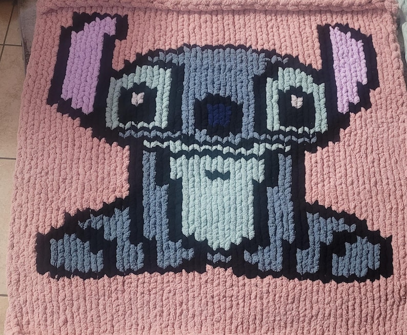May include: A pink blanket with a large, pixelated cartoon character design. The character is depicted in shades of grey, black, and blue, with pink ears. The blanket is made from a thick, textured yarn, suitable for a child's bedroom or a cosy living area.