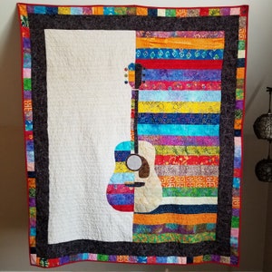 May include: A colourful quilt featuring a guitar design. The quilt is made from a variety of fabrics in shades of red, blue, green, yellow and orange. The guitar is outlined in black and has a white body. The quilt is framed with a black border and a red, white and blue trim.