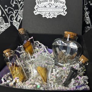 May include: A black box labelled "House of Hexes, Heirlooms & Hypnotics" contains glass bottles with cork stoppers. The bottles hold a butterfly wing, dried herbs, and a small octopus in a heart-shaped bottle. The box is filled with shredded paper.