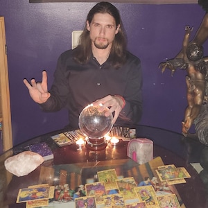 May include: A round glass table covered in a spread of tarot cards, a clear crystal ball on a wooden stand, and two small candles. A person with long dark hair sits behind the table, wearing a black button-down shirt, with a large bronze angel statue nearby.