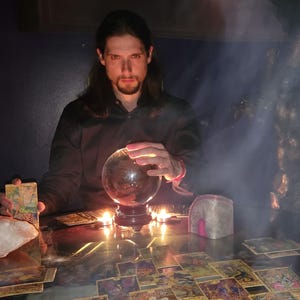 Könnte beinhalten: Eine Person mit langen dunklen Haaren sitzt an einem Tisch mit Tarotkarten, einer Kristallkugel und Kerzen. Die Szene ist schwach beleuchtet und hat eine mystische Atmosphäre. Eine große Statue befindet sich im Hintergrund, und Rauch trägt zum geheimnisvollen Ambiente bei.