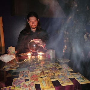 Psychic Derek conducting a spiritual reading with a framed Virgin Mary and Child picture on the wall above a candlelit tarot table adorned with crystals.