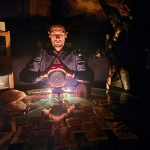 May include: A clear glass crystal ball sits on a dark stand, illuminated from within, positioned on a glass table covered with a spread of rectangular tarot cards. A rough-textured, light-coloured stone sits to the left of the cards.