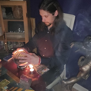 Psychic Derek offering deep energy healing and intuitive therapy, seated near drawn tarot cards, natural crystal clusters, and a clear crystal sphere.
