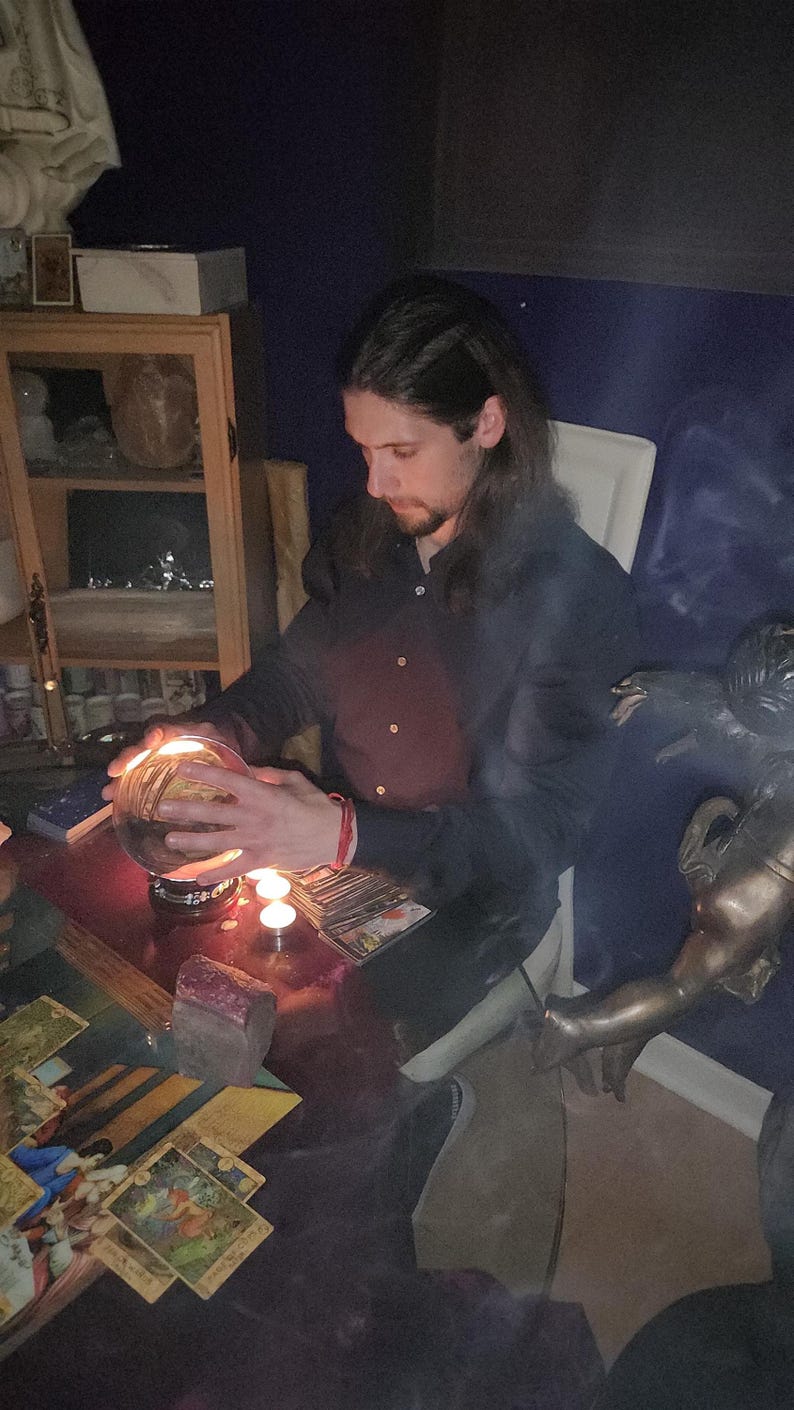 Psychic Derek offering deep energy healing and intuitive therapy, seated near drawn tarot cards, natural crystal clusters, and a clear crystal sphere.