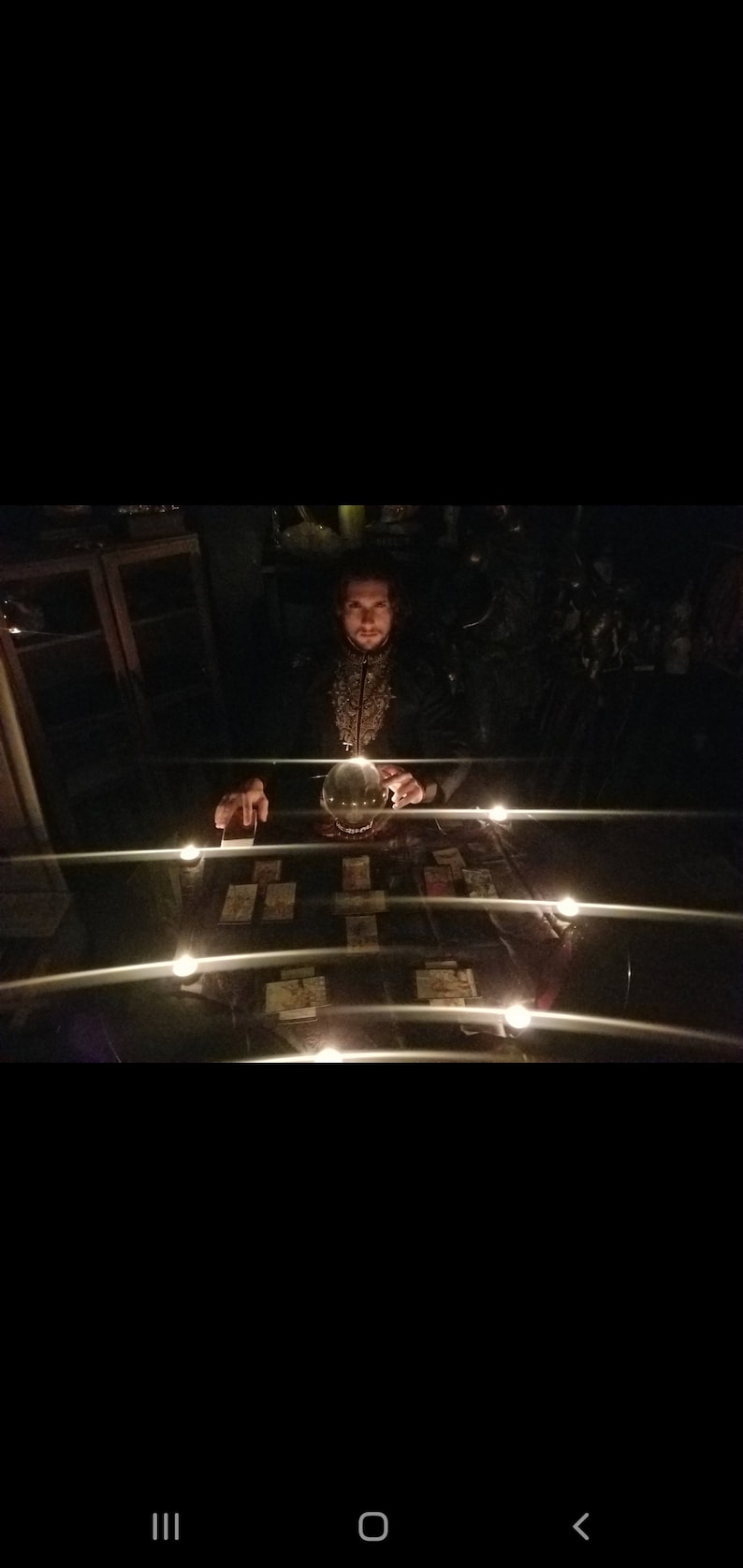 May include: A man in a dark suit sits at a table with a crystal ball and tarot cards. The table is lit by candles and the room is dark.