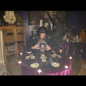 May include: A person sits at a table with a purple tablecloth and a glass top. They are holding a crystal ball in their hands. There are tarot cards spread out on the table and candles around the edge of the table.