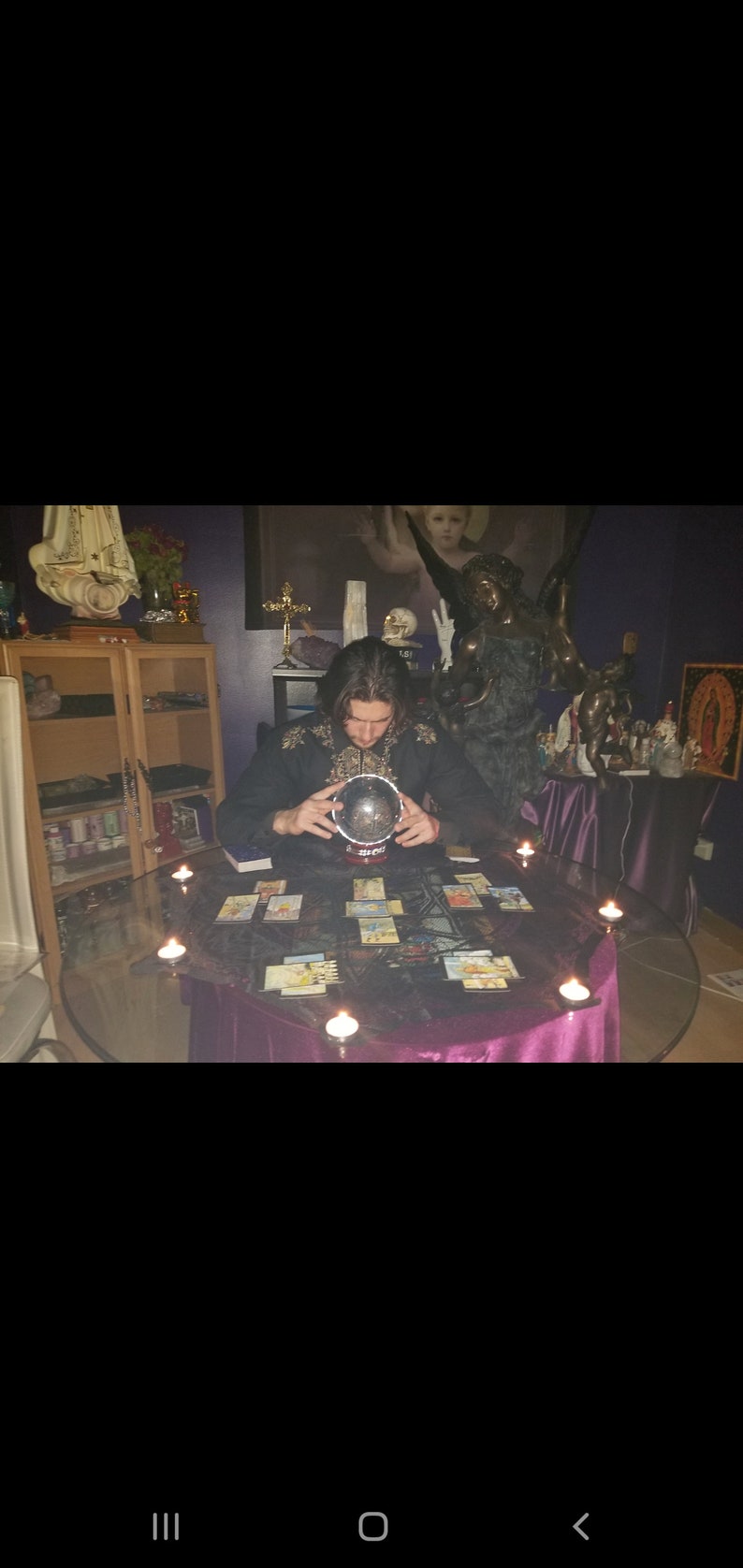 May include: A person sits at a table with a purple tablecloth and a glass top. They are holding a crystal ball in their hands. There are tarot cards spread out on the table and candles around the edge of the table.