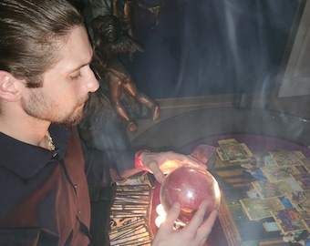 May include: A clear glass sphere illuminated from beneath by a warm light source, held by two hands above a dark surface covered in numerous rectangular tarot cards. Wisps of white smoke drift upward around the sphere and the cards.