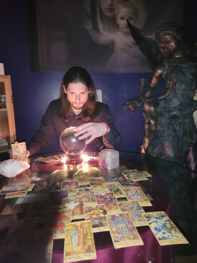Intuitive advisor Psychic Derek channeling higher divine energy near a carved angel statue, drawn tarot cards, polished stones, and lit pillar candles.