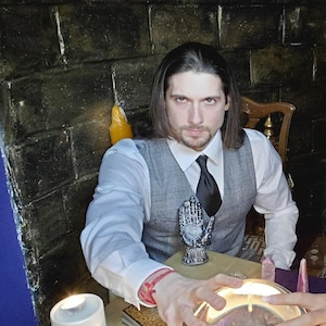 May include: A person wearing a white dress shirt, a grey plaid vest, and a black necktie. On the table sits a clear crystal ball, a white pillar candle with a religious image, a small decorative hand sculpture, and a deck of tarot cards.