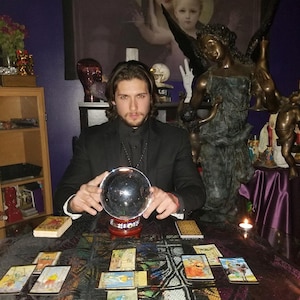 Intuitive advisor Psychic Derek channeling higher divine energy near a carved angel statue, drawn tarot cards, polished stones, and lit pillar candles.