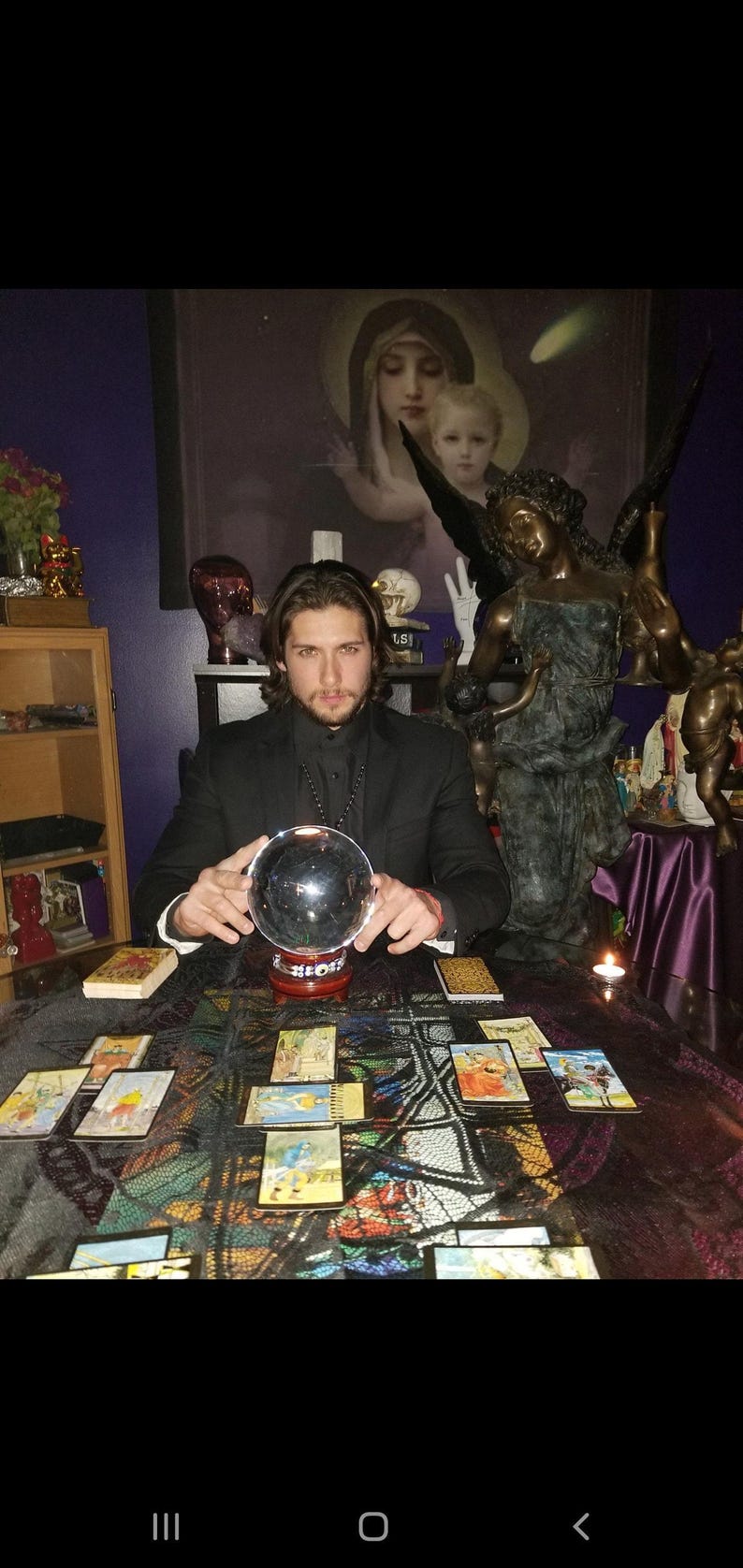 Intuitive advisor Psychic Derek channeling higher divine energy near a carved angel statue, drawn tarot cards, polished stones, and lit pillar candles.