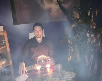 May include: A person in a black button-down shirt sits at a glass table covered in numerous tarot cards. A glowing crystal ball rests on a black stand in the centre of the table. A large bronze-coloured angel statue stands to the right of the table.