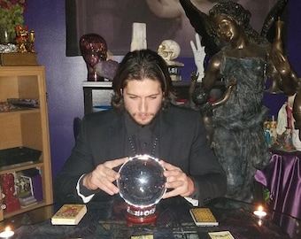 May include: A clear glass crystal ball sits on a dark red base, positioned on a table covered with a patterned cloth and multiple tarot cards. The sphere reflects light, and the scene includes a dark-suited figure and a bronze-colored angel statue.