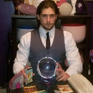 Psychic Derek offering deep energy healing and intuitive therapy, seated near drawn tarot cards, natural crystal clusters, and a clear crystal sphere.