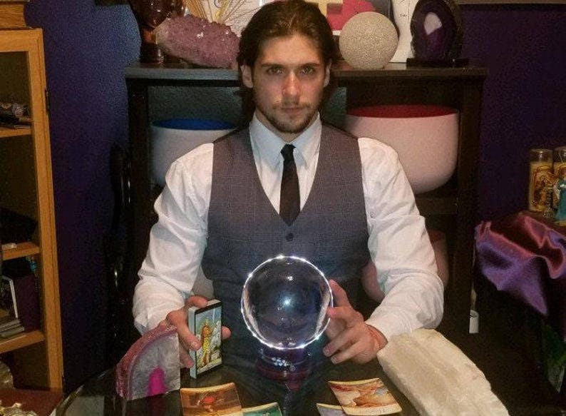 Psychic Derek offering deep energy healing and intuitive therapy, seated near drawn tarot cards, natural crystal clusters, and a clear crystal sphere.