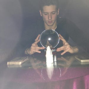 Psychic Derek conducting an online psychic reading beside a serene angelic figurine, natural gemstone crystals, a crystal ball, and burning incense smoke.