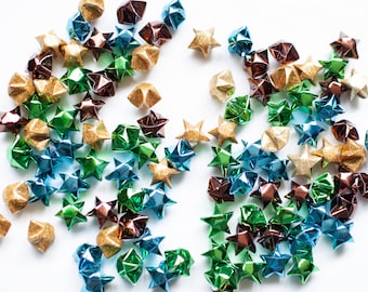 Puede incluir: Una colección de estrellas de origami en varios colores, incluyendo dorado, verde, azul y marrón. Las estrellas están hechas de papel metálico y están esparcidas sobre un fondo blanco.