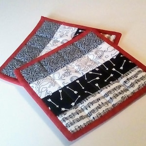 Puede incluir: Dos agarraderas de olla acolchadas con ribete rojo. Una agarradera tiene un patrón en blanco y negro con notas musicales. La otra agarradera tiene un patrón en blanco y negro con flechas y diseños florales.