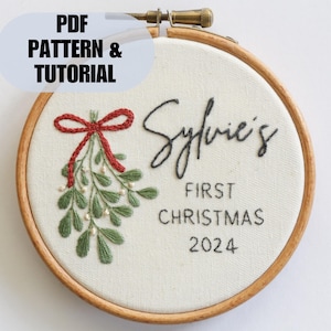 Pode incluir: Aro bordado com um laço vermelho, folhas verdes e contas brancas. O texto "Sylvie's First Christmas 2024" está bordado em fio preto.