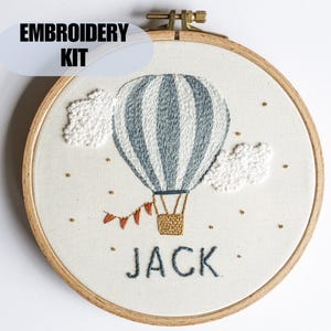 May include: An embroidery kit featuring a hot air balloon design. The balloon is blue and white striped, with a brown basket and small orange flags. The name "JACK" is embroidered below the balloon. The text "EMBROIDERY KIT" is above the design.
