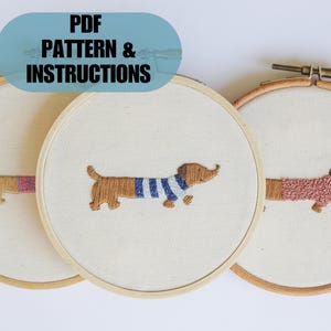 May include: Three embroidery hoops with different dachshund designs. The hoops are made of wood and have a natural finish. The dachshunds are embroidered in brown, pink, and blue thread. The text "PDF PATTERN & INSTRUCTIONS" is displayed in a blue box.