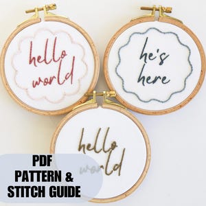 Op de afbeelding: Drie borduurringen met houten frames. Twee ringen hebben de woorden "hello world" in rode en gouden draad, en één heeft "he's here" in blauwe draad. De ringen staan op een witte achtergrond. Een blauw ovaal aan de onderkant luidt "PDF PATTERN & STITCH GUIDE."