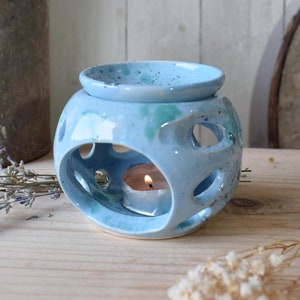 Op de afbeelding: Een lichtblauwe keramische olieverdamper met een rond, open ontwerp en een kleine, witte theelichtkaars erin. De diffuser heeft een gespikkelde glazuur en een klein, rond schoteltje bovenop voor etherische oliën.