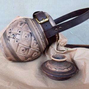 Könnte beinhalten: Ein handgefertigtes, hellbraunes Keramikgefäß mit einem dunkelbraunen Ledergriff und einer Schnalle. Das Gefäß weist geschnitzte geometrische Muster und einen passenden Deckel auf. Der Griff ist mit Messingbeschlägen befestigt. Der Deckel liegt neben dem Gefäß auf braunem Papier.