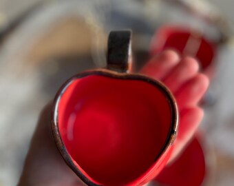 Könnte beinhalten: Eine rote herzförmige Keramiktasse mit einem braunen Henkel. Die Tasse wird in einer Hand gehalten, und das Innere der Tasse ist sichtbar.